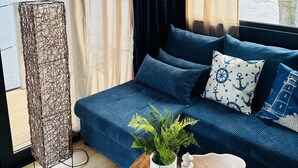 A modern living room with a blue sofa, nautical-themed pillows, a wooden coffee table, and a view of a river and buildings. - Premium Hausboot Blaue Lagune (Lübeck)