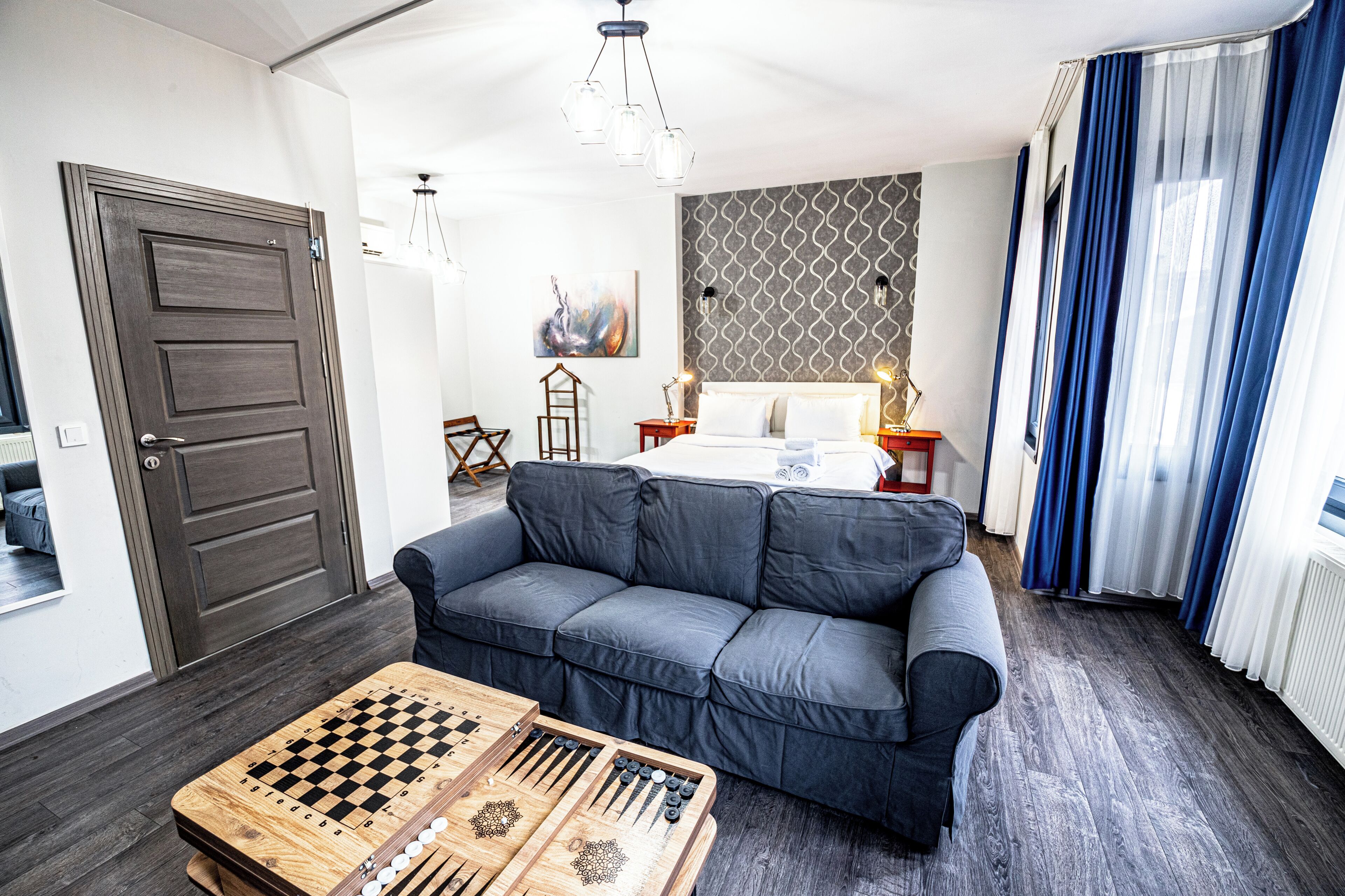 Habitación de hotel moderna con sofá gris, una mesa de juego de ajedrez de madera, cama con ropa blanca y pared con estampado.