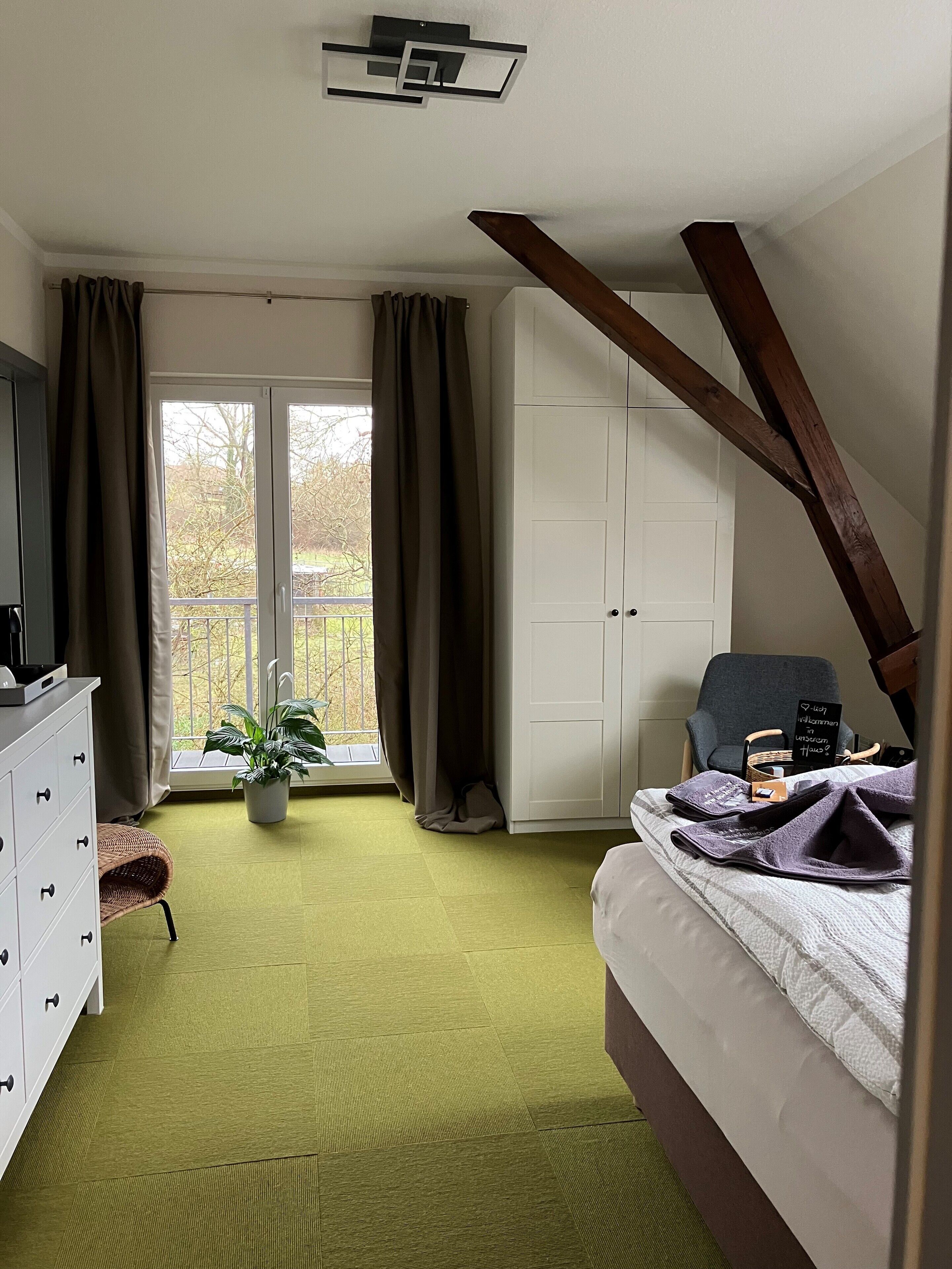 A bedroom with a bed, a dresser, a chair, a potted plant, and a view of the outdoors through a large window.