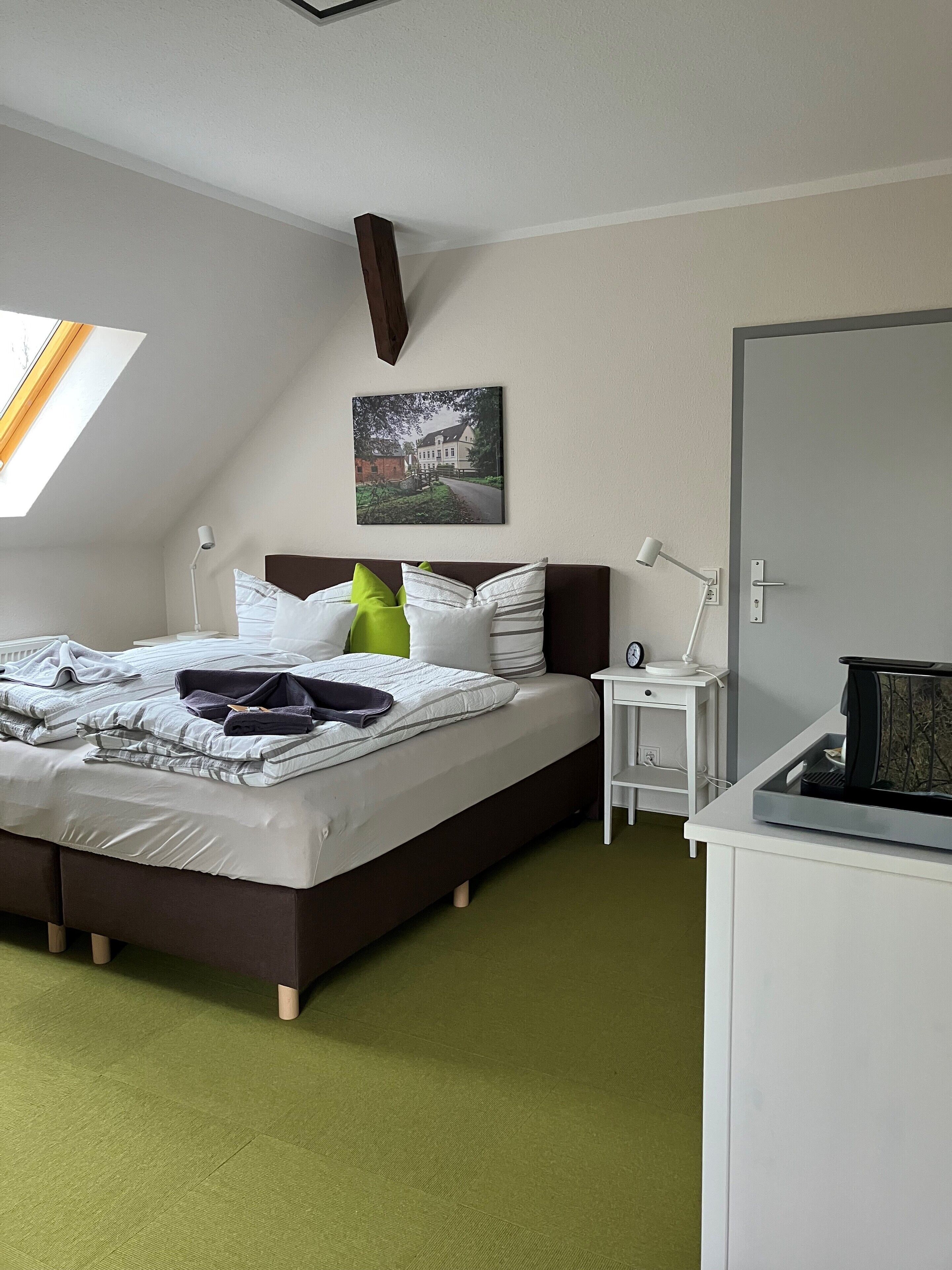 Cama bem arrumada com almofadas e cobertor, mesa de cabeceira com abajur, relógio e torradeira. Um quadro pendurado na parede acima da cama.