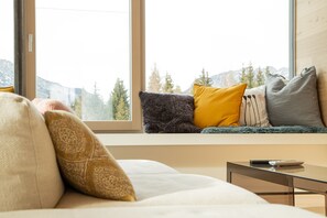 Un coin salon confortable avec un canapé et des coussins, une petite table et une fenêtre donnant sur un paysage de montagnes et d’arbres.
