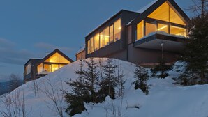 Une maison moderne à deux étages, dotée de grandes fenêtres et d’un toit en pente, entourée de neige et d’arbres.