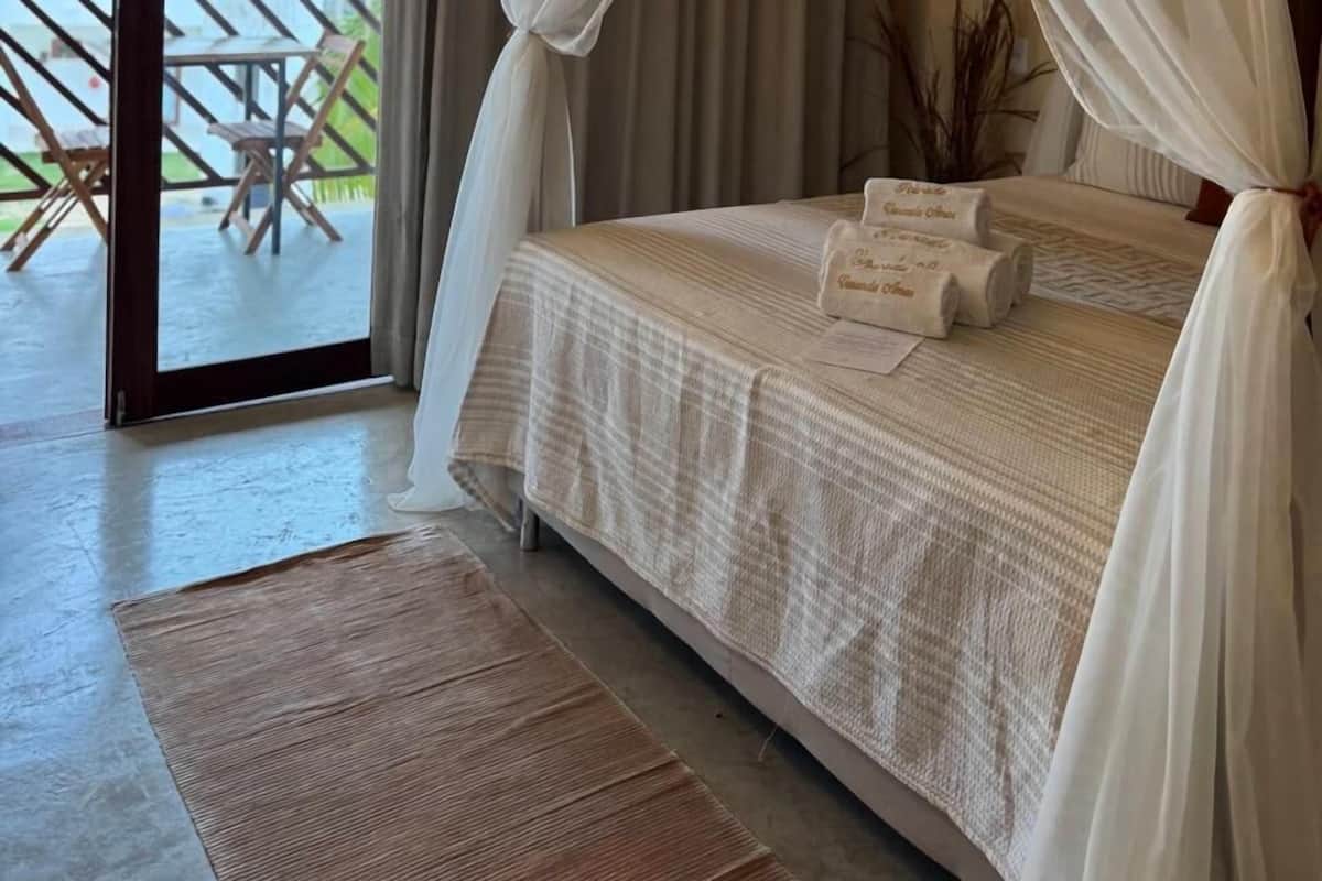 Une chambre à coucher avec un lit, des rideaux blancs et une vue sur un balcon aménagé avec du mobilier d’extérieur.
