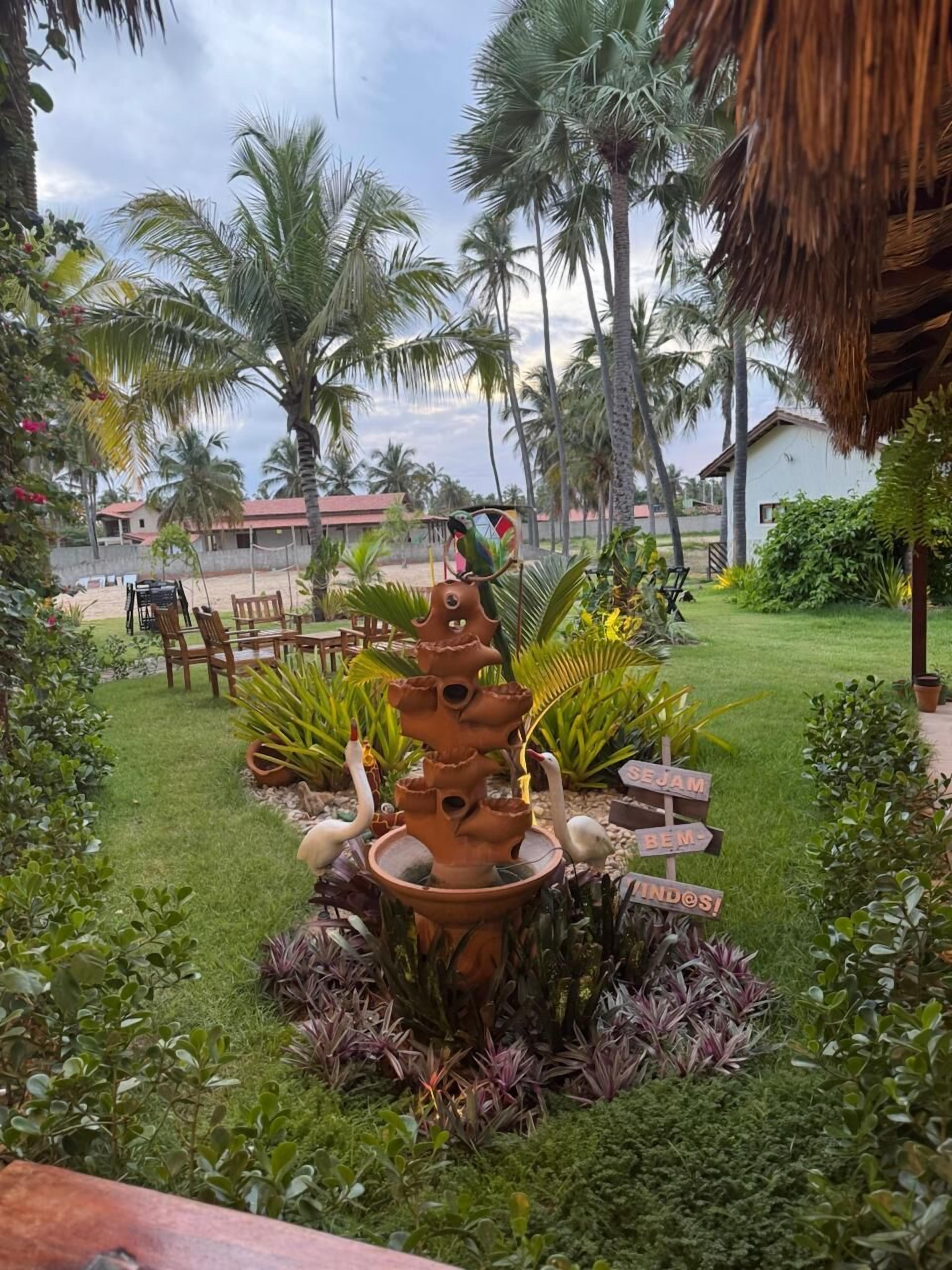 A tropical garden with a bird fountain, palm trees, and a thatched-roof structure.