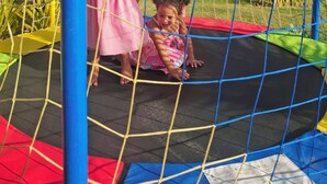 Children playing on a trampoline with a netted enclosure, surrounded by greenery and a building with a thatched roof. - Pousada Varanda Amar (Amontada)