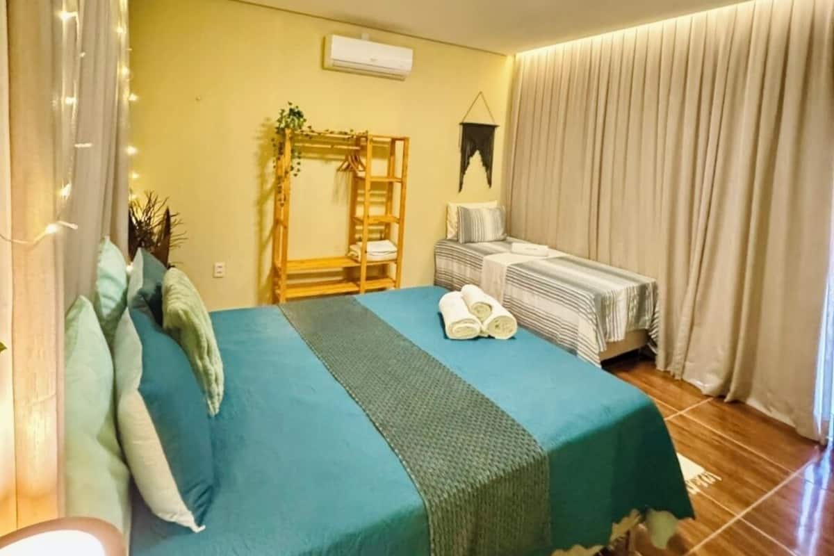 Une chambre d’hôtel avec un lit, une table de chevet, une lampe, une plante suspendue et une fenêtre avec des rideaux.