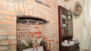 A brick fireplace with a painting of a rabbit and flowers, a wooden cabinet with dishes, and a mirror on the wall. - Oakman Cottage (Honiton)