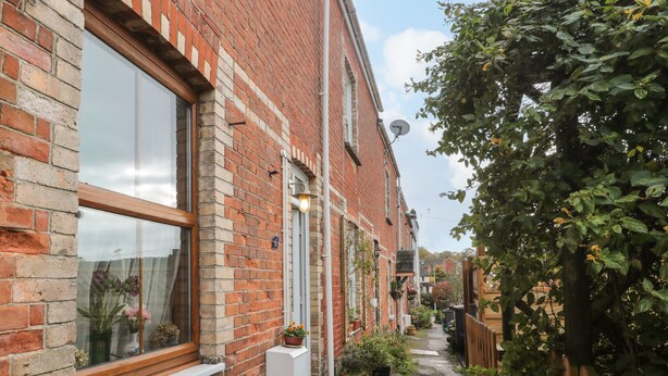 A narrow alleyway with a brick building on one side and a wooden fence on the other. There are potted plants along the alley and a small garden area. - Oakman Cottage (Honiton)