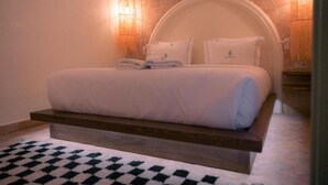 A neatly made bed with a headboard, a wooden bedside table, and a checkered rug on the floor. - Riad Lalla Mimouna - Suite (Marrakech)