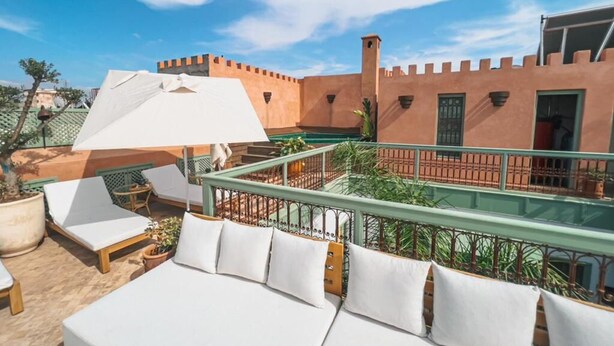 A rooftop terrace with white lounge chairs and a covered seating area, overlooking a traditional building with a red-tiled roof and a green railing. - Riad Lalla Mimouna - Suite (Marrakech)