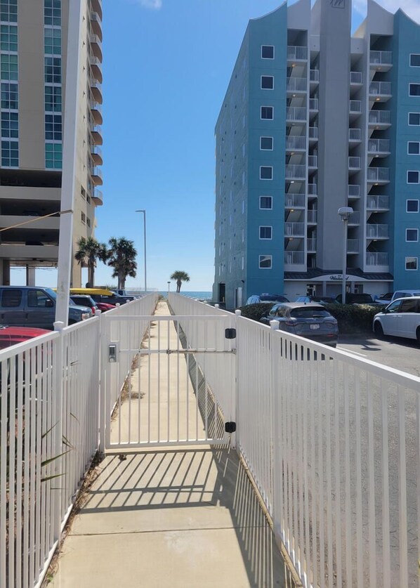 A pedestrian walkway with a white fence, leading to a parking area with cars and a building with balconies. - New Listing! Luxury Home, Sleeps 10 w/ Shared Pool (Gulf Shores)