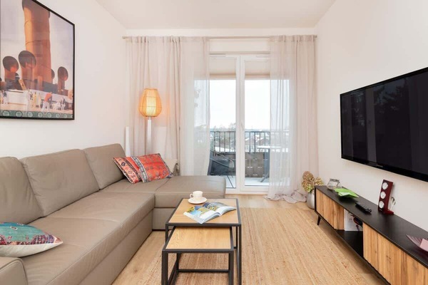 A bright living room with a beige sofa, a wooden coffee table, a TV, and a large window with white curtains.