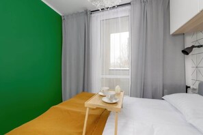 A hotel room with a bed, a wooden bedside table with two cups and a tray, a green wall, and a window with grey curtains. - Bright Apartment with Bedroom | Good Location in Katowice. (Katowice)