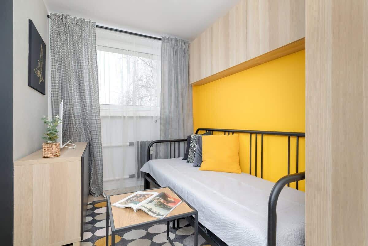 A compact bedroom with a bed, a bedside table with a magazine, a small cabinet, and a window with curtains.