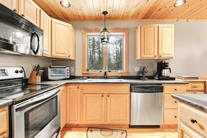 A kitchen with wooden cabinets, stainless steel appliances, a window with a view of trees, and a hanging light fixture. - Deer Run - Chalet w/ Fireplace (Greenwood)
