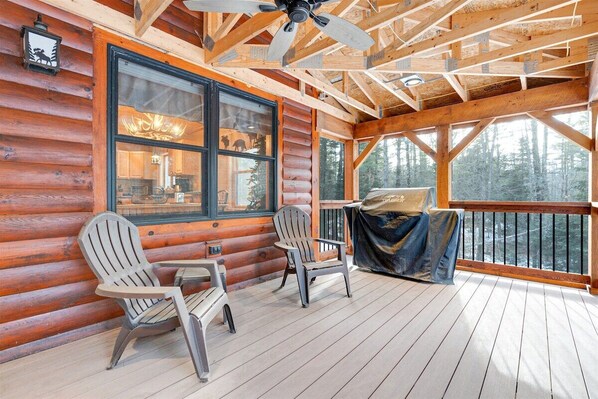 A log cabin with a wooden deck, two Adirondack chairs, and a large window overlooking a forest.