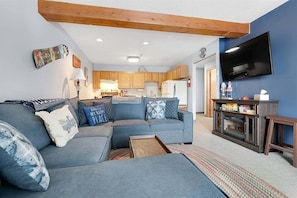 A living room with a sectional sofa, a television, and a kitchen in the background. - White Cap B-113 - Sunday River Condo (Newry)