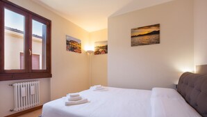 A neatly made bed with white linens and towels, a window with a view of a building, and two wall-mounted pictures. - Apartment Bella Sathea (Torri del Benaco)
