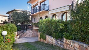 A two-story building with a stone facade, a balcony with a black railing, and a stone wall with a small garden area. - Apartment Bella Sathea (Torri del Benaco)
