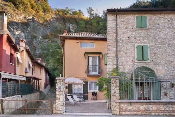 A narrow street with stone buildings, a stone gate, and a small outdoor seating area with a table and chairs.