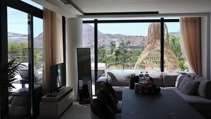 Ein modernes Wohnzimmer mit Flachbildfernseher, einer Couch, einem Esstisch und großen Fenstern mit Blick auf eine Berglandschaft.