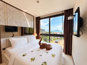 A hotel room with a bed, a television mounted on the wall, and a view of the city through large windows. - BO202- 1 Bedroom Serviced Apartment With Sea View at Ao Nang Beach (Tambon Ao Nang)
