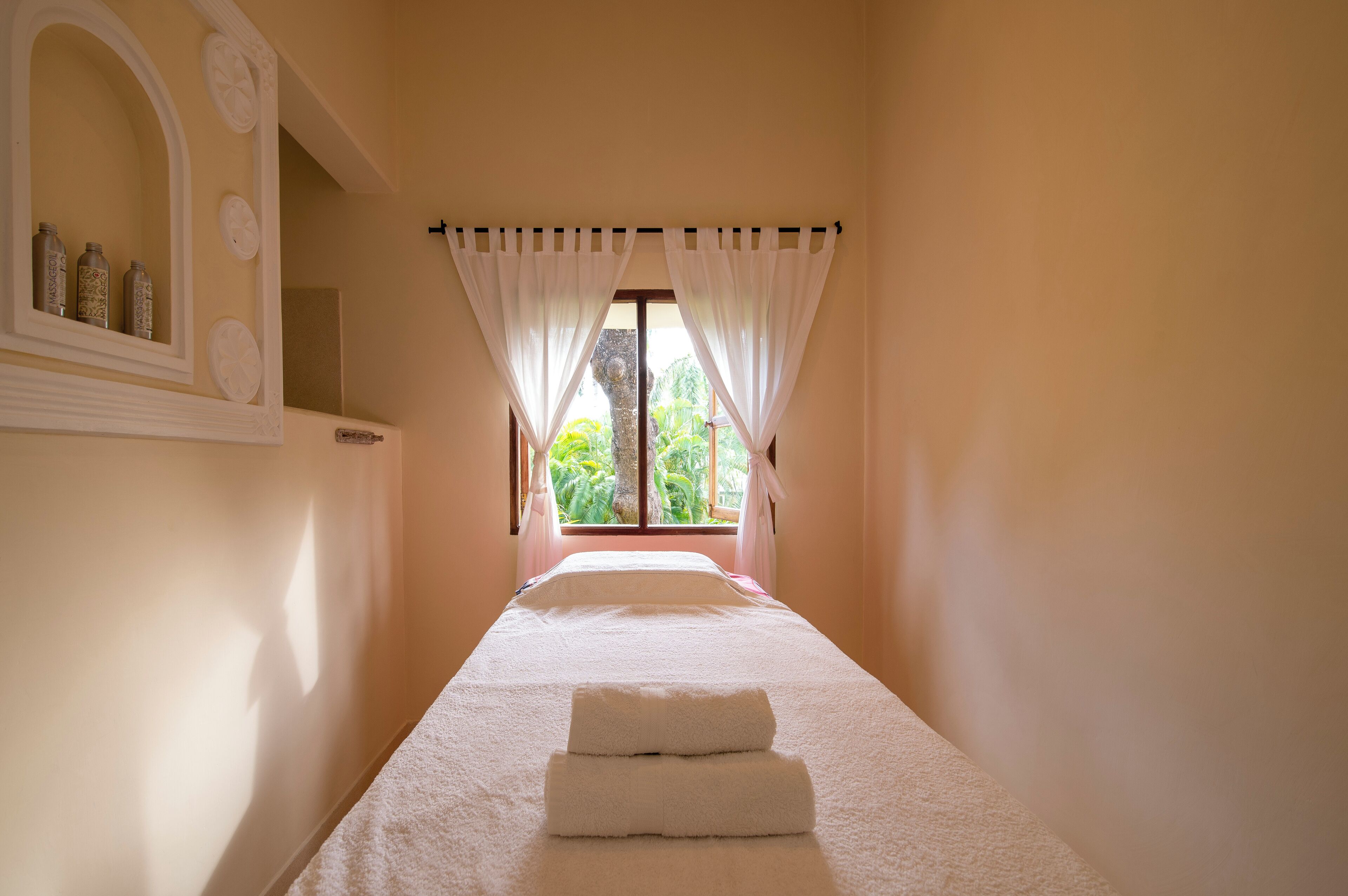 A narrow room with a bed, a window with curtains, and a mirror with decorative items.