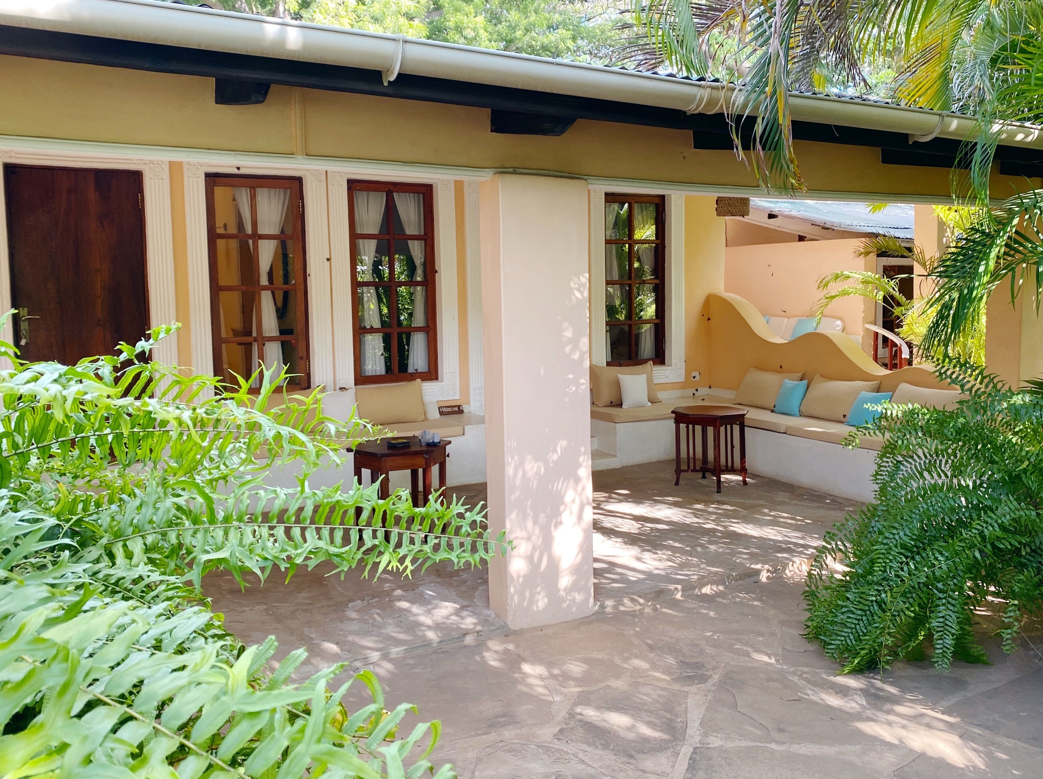 A covered outdoor seating area with a sofa and a small table, surrounded by lush greenery.