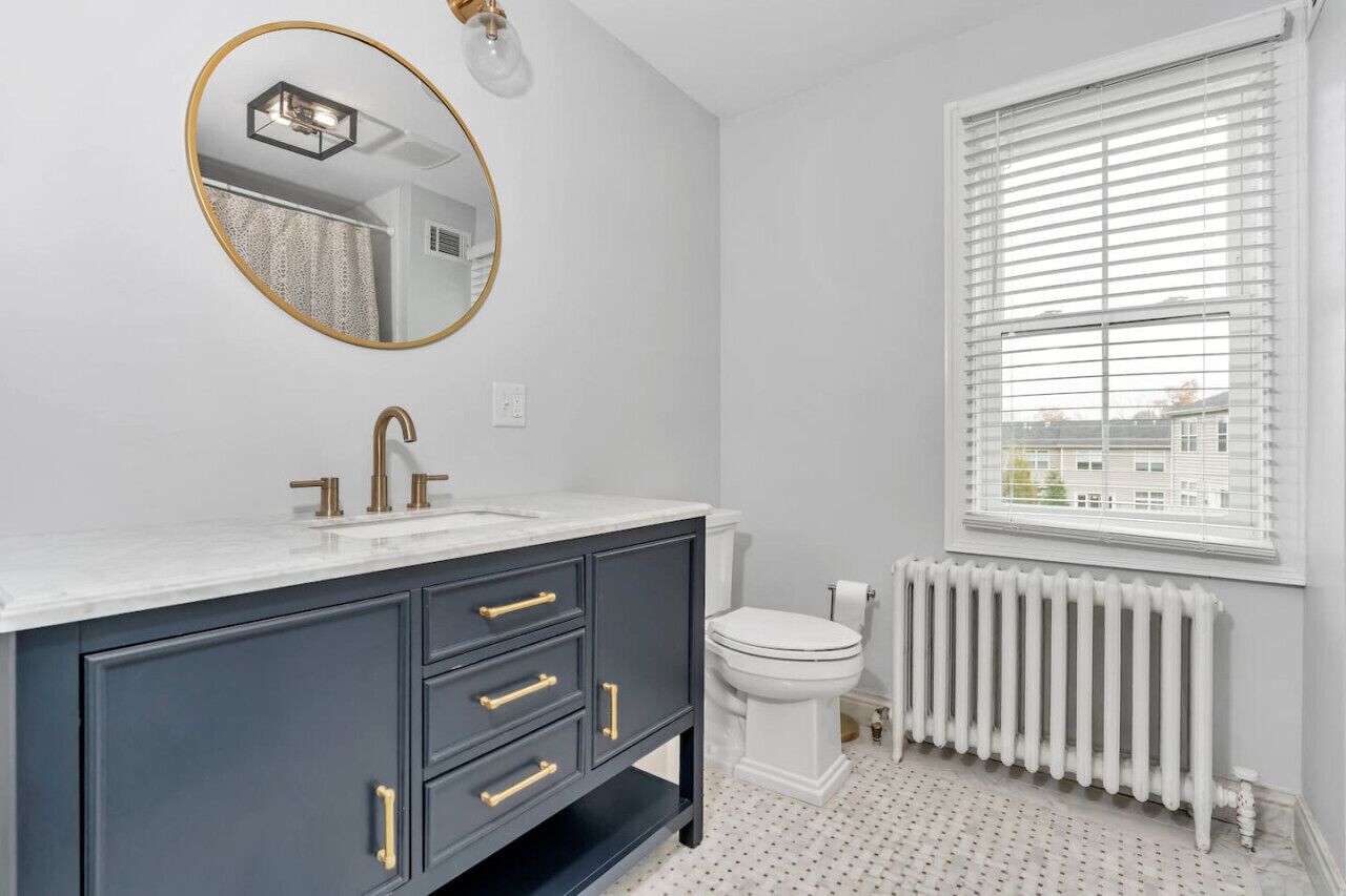 A bathroom with a white vanity, gold fixtures, a round mirror, a toilet, and a window with blinds.