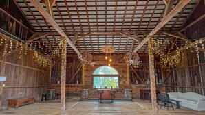 A rustic barn interior with wooden beams, a large arched window, and string lights.