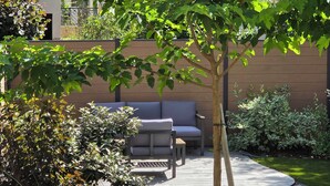 Eine Terrasse mit einer Couch und einem Tisch, umgeben von Grünpflanzen und einem Holzzaun.