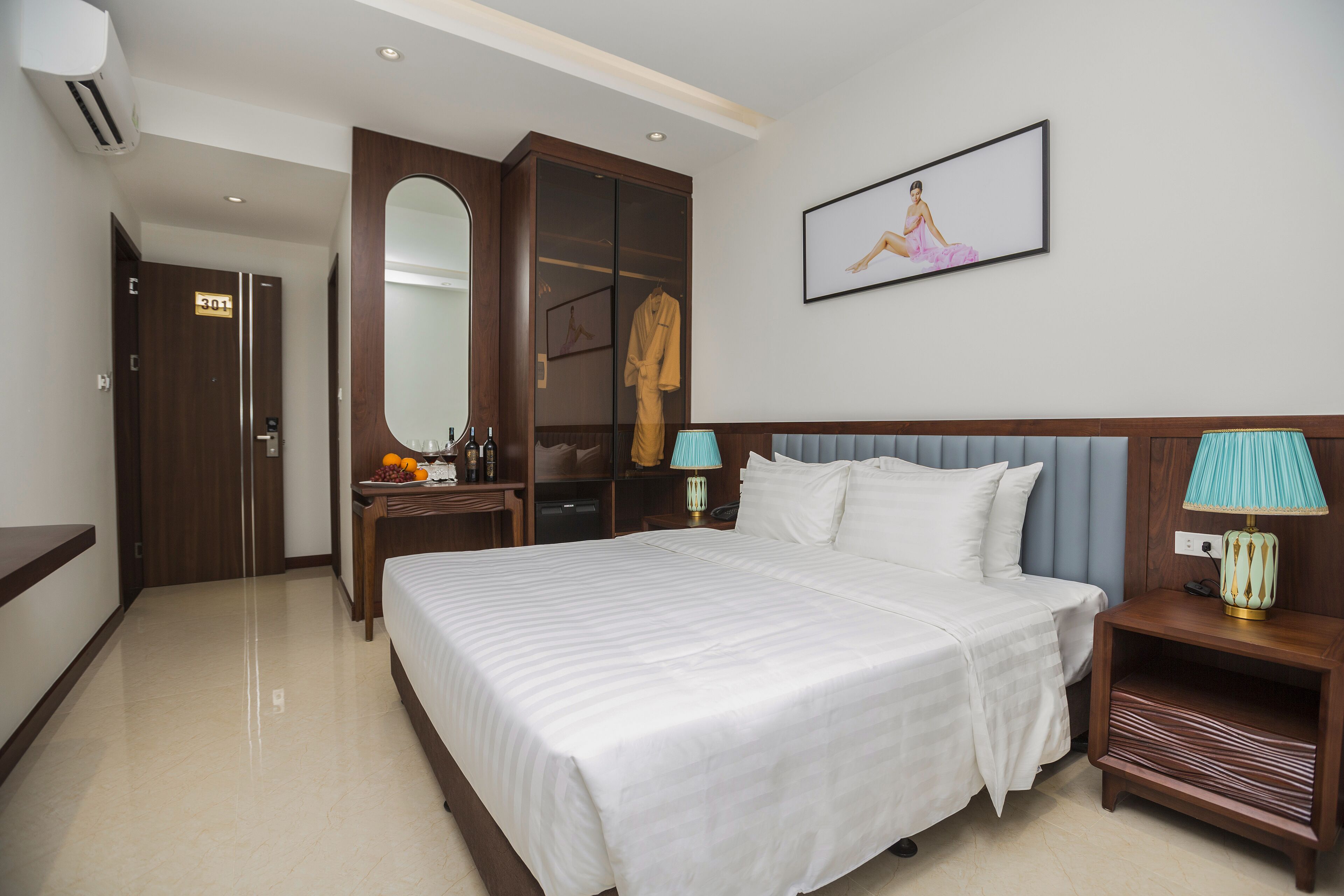 Quarto de hotel com uma cama grande, um guarda-roupa, um espelho, uma mesa de cabeceira com uma lâmpada e um quadro na parede.