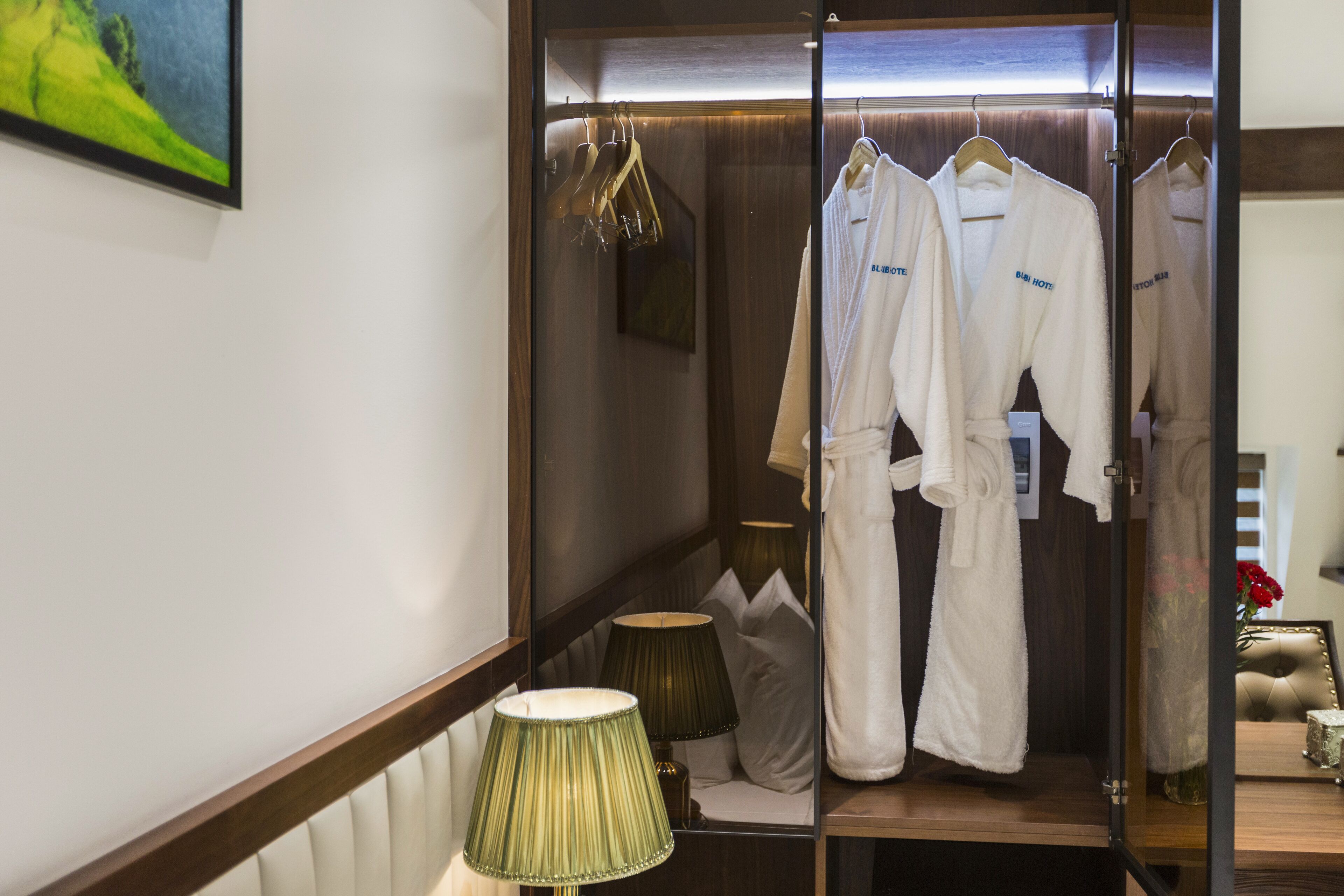 A hotel room with a mirror, a green lamp, a wooden vanity, and white robes hanging on a rack.