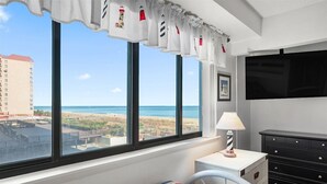 Quarto de hotel com uma janela grande, uma cama com colcha listrada, uma mesa de cabeceira com abajur e um aparador.