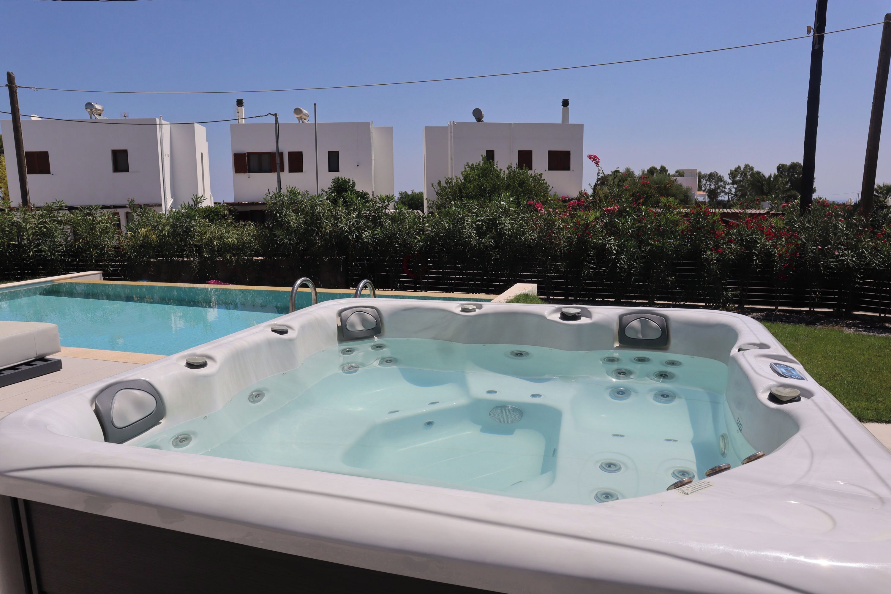 A hot tub with a clear pool in the background, surrounded by a fence and greenery.