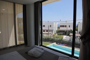 A hotel room with a large window, a bed with white linens, and a view of a swimming pool and buildings outside. - Trapezia Luxury Living - Morganite (Afantou)
