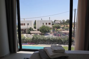 A bedroom with a large window, a bed, and a view of a swimming pool and buildings.