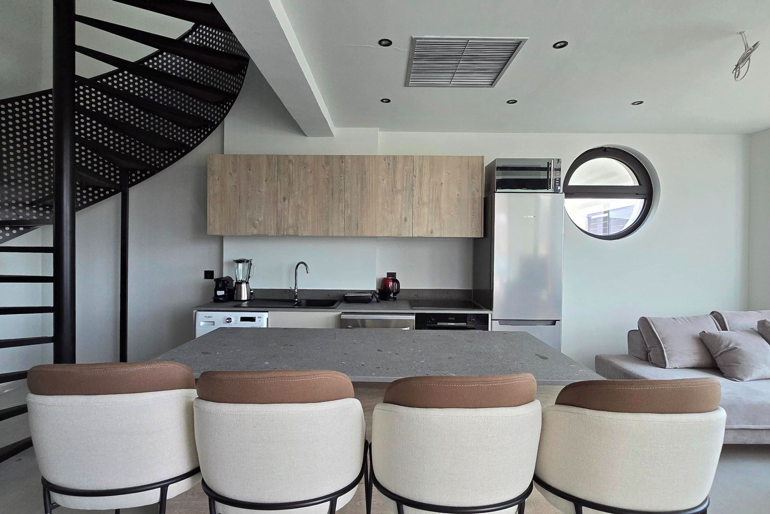 A modern kitchen with a central island, a dining area with four chairs, a staircase, and a round window.