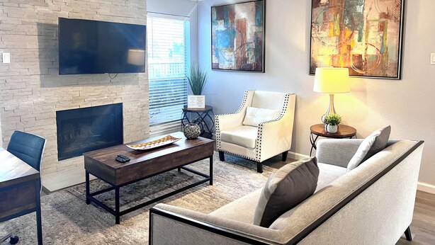 A living room with a fireplace, a ceiling fan, a flat-screen TV, a coffee table, a sofa, and a lamp. - I20 - Gated 2BD Condo | Irving | DFW 10 Min | Pool (Irving)