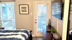 A bedroom with a bed, a door with blinds, a framed picture on the wall, and a small table with a plant. - I20 - Gated 2BD Condo | Irving | DFW 10 Min | Pool (Irving)