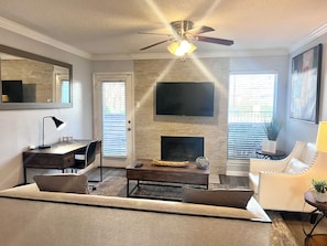 A living room with a fireplace, a ceiling fan, a TV mounted on the wall, and a seating area with a sofa and a chair.