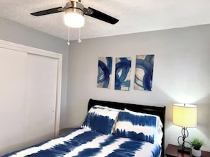 A bedroom with a bed, a ceiling fan, a nightstand with a lamp, and abstract wall art.