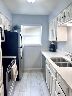 A compact kitchen with white cabinetry, a black refrigerator, a built-in oven, a double sink, and a window with blinds.