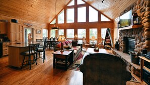 A spacious living room with a high ceiling, wooden floors, a fireplace, and large windows. - Twin Pines: Dog-Friendly Lakefront Cabin w/ Private Dock & Kayaks (Stone Lake)