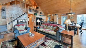 A spacious log cabin interior with a high ceiling, a staircase leading to an upper level, a cozy living area with a sofa and coffee table, and a kitchen area. - Twin Pines: Dog-Friendly Lakefront Cabin w/ Private Dock & Kayaks (Stone Lake)
