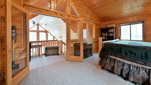 A log cabin bedroom with a lofted sleeping area, a bookshelf, and a large window. - Twin Pines: Dog-Friendly Lakefront Cabin w/ Private Dock & Kayaks (Stone Lake)