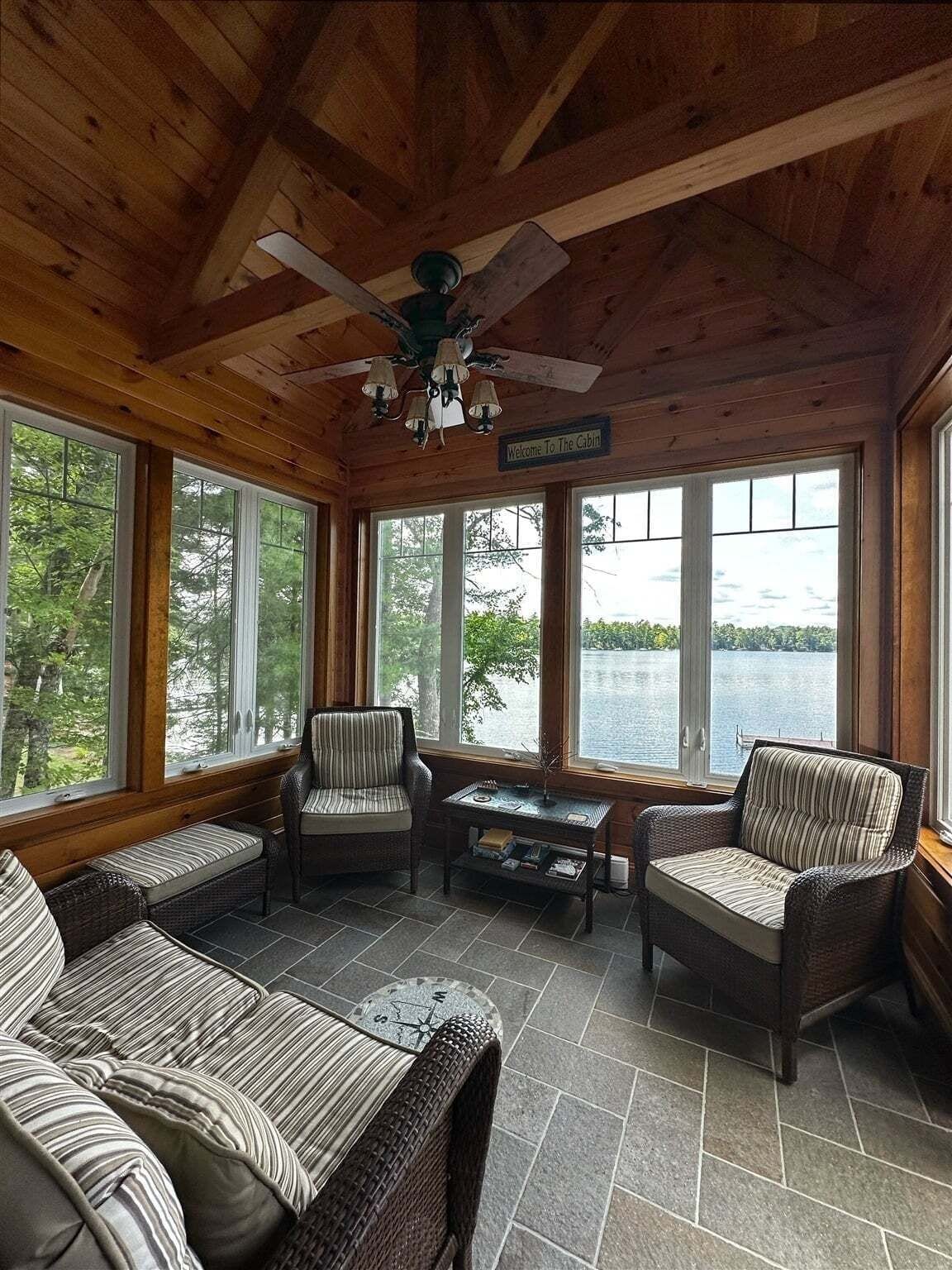 A cabin with a wooden interior, large windows, a ceiling fan, and a view of a lake.