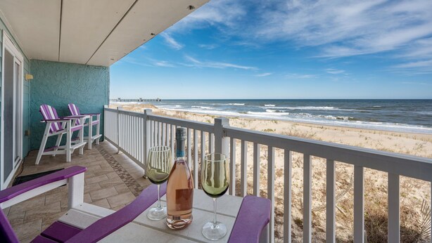 Balkon med udsigt til stranden, to stole, et bord med en flaske og glas, samt gelænder.