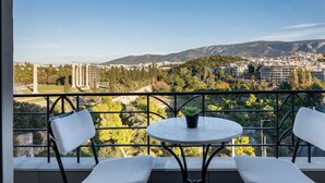 Un balcón con una mesa redonda y dos sillas blancas, con vista a un paisaje urbano que incluye edificios y montañas.