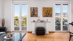 Amplio salón con una mesa de centro de vidrio, un sofá de cuero negro y una chimenea blanca. Hay dos ventanas grandes con vistas a la ciudad y dos cuadros en la pared.